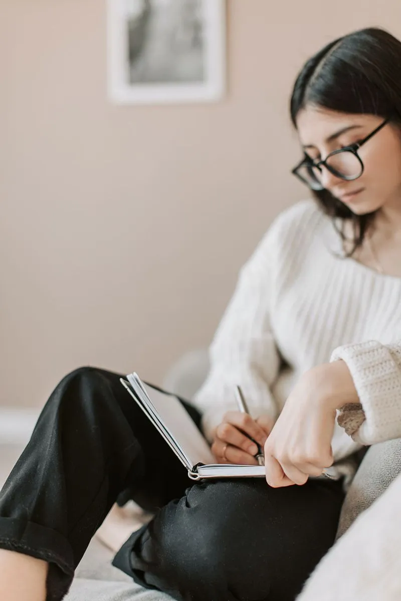 Young woman with glasses in a white sweater writing in a journal on a bed